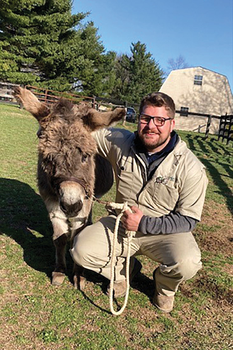 A vet kneels to pose for a photo with a mini donkey