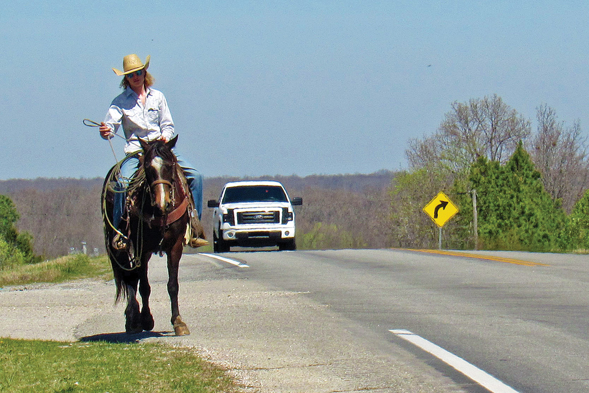 A cowgirl riding her horse along a road with a truck driving behind them