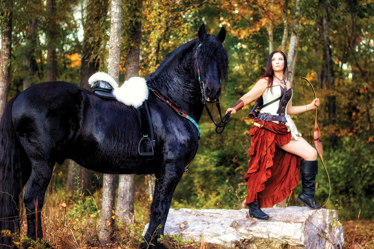 A girl in an archery costume poses with a large black horse
