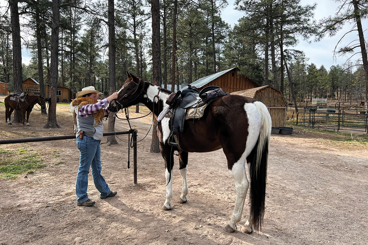A trail guide with a cat on her shoulder pats a pinto that's tacked up and ready for a ride