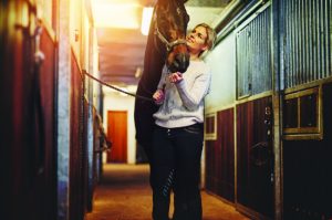 Smiling young woman preparing her chestnut horse for a ride while standing inside a stable on a farm.